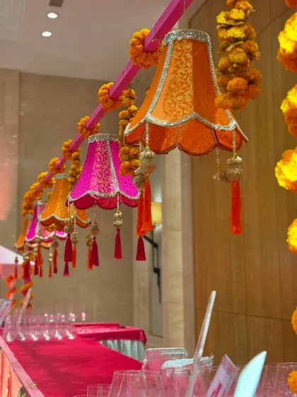 A row of custom-made fabric lampshades in shades of pink and orange, hanging above a service counter. These were embellished with golden tassels and strung together with marigold garlands for a festive Garba night.