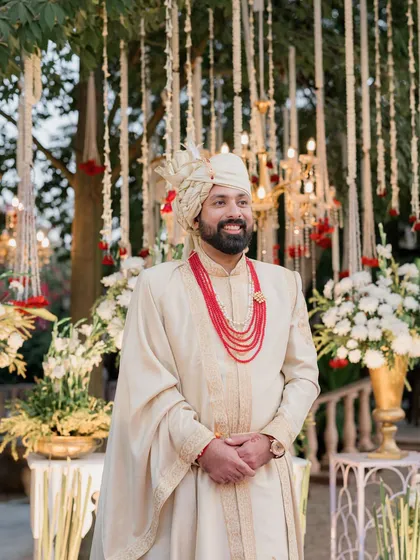 A smiling groom stands before a backdrop of hanging floral strings and chandeliers at his Lohagarh Fort wedding. The decor blends traditional elements with opulent touches.