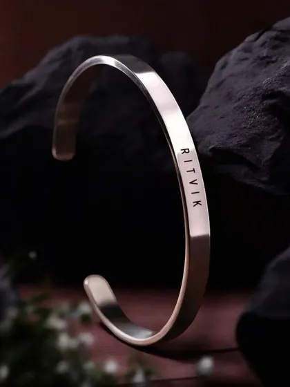A silver-finish cuff bracelet engraved with 'RITVIK', displayed against a dark, textured rock. This highlights the contrast and clean lines of the engraving.