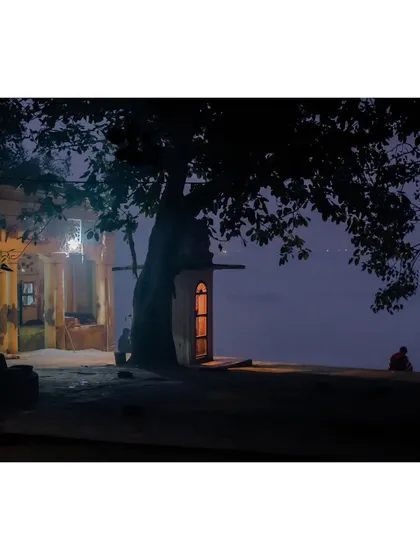 A serene night scene on the banks of the Yamuna. A small, illuminated shrine sits under a large tree, while a lone figure sits by the water's edge in quiet contemplation.