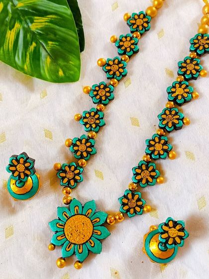 This pretty floral necklace set is shown in a sea-green and yellow combination. The chain is made of small flowers, leading to a larger central flower, and includes matching jhumkas.