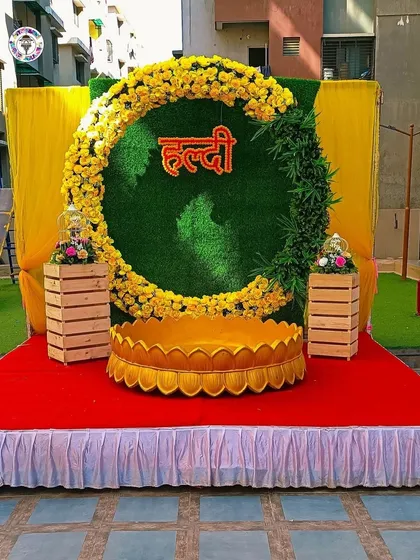 This Haldi decor features a large circular frame against a green backdrop, with the word "Haldi" in Hindi at the center. The crescent-shaped floral arrangement is made of bright yellow marigolds.