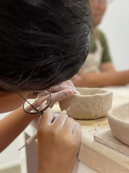 A student uses a needle tool to incise a small, personal mark onto their hand-built bowl.