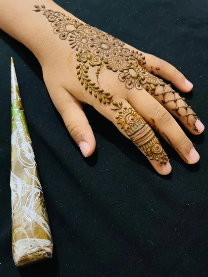 A delicate Arabic style design on a client's hand, shown next to the henna cone used to create it.