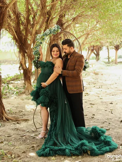 The couple poses by a birdcage prop in the woods. The rich green of her dress and the natural surroundings create a magical, forest-like atmosphere for their maternity shoot.