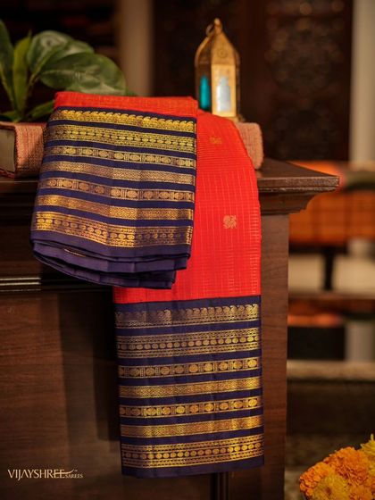 A timeless red Kanjivaram with a deep blue contrast border, featuring multiple bands of traditional patterns woven in gold zari. This piece was personally sourced by our proprietor.