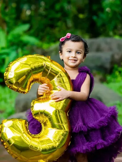A proud three-year-old girl holds a large golden '3' balloon during her outdoor birthday photoshoot.