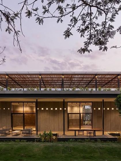 The building's facade at dusk, framed by native trees. The combination of the thatched upper roof, large glass openings, and warm interior lighting creates a sense of transparency, blurring the boundary between the indoor spaces and the surrounding garden.