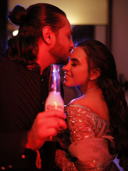 An intimate, candid shot from an engagement party. The groom kisses the bride's forehead under a warm red light, creating a romantic and moody photograph.