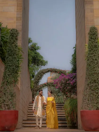 The couple walks down a beautifully landscaped staircase, hand in hand, creating a grand and romantic frame from their Haldi day.