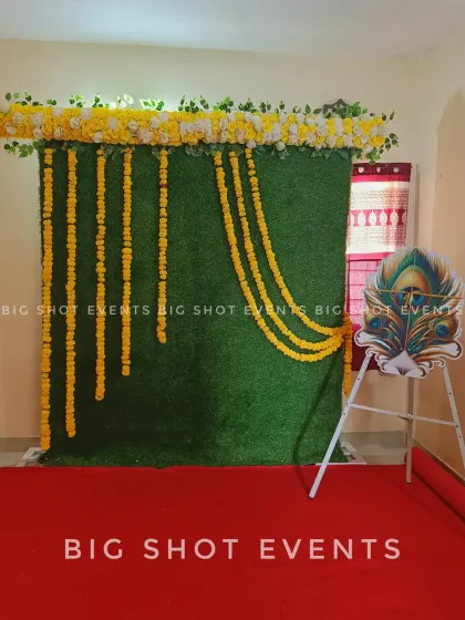 A clean and simple backdrop for a cradle or naming ceremony. This setup uses a green turf wall decorated with marigold garlands and a floral top border. A peacock feather cutout on an easel adds a nice decorative touch.
