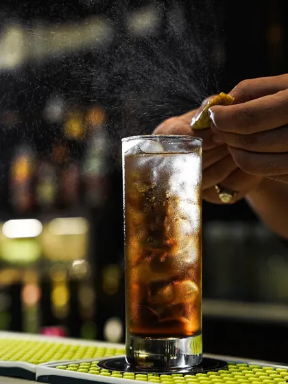A bartender's hands captured in motion, spritzing a lemon zest over a classic long island iced tea. The focus on the action highlights the craft and care that goes into making a perfect cocktail.