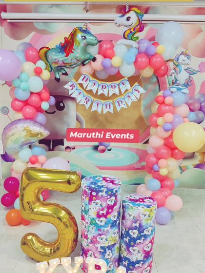 A unicorn party setup with a pastel rainbow backdrop. The decor includes unicorn foil balloons, a large number "5" balloon, and themed stands.