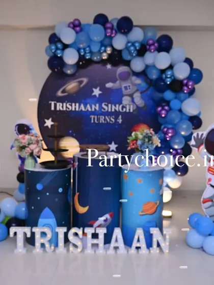 An out-of-this-world space theme for a 4th birthday party. The decoration includes a galaxy backdrop, themed pedestals, cutouts of an astronaut and a rocket ship, and a blue and purple balloon arch to create a cosmic adventure.