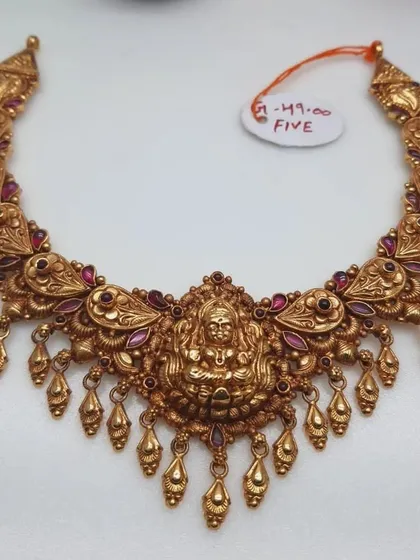 This intricate temple necklace features a central goddess motif and is adorned with ruby-colored stones and golden bead drops. It is made from 92.5 silver with gold plating.