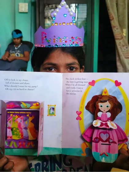 A child proudly displays a pop-up book and a crown she made during a Story Sparkle activity. For many children, stories of princesses are not just tales, but a discovery of their own bravery and resilience.