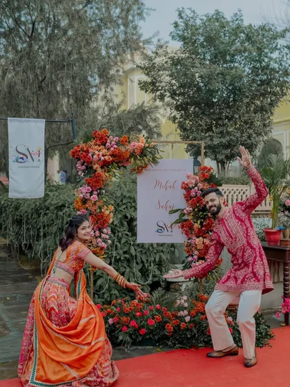 The couple sharing a playful moment at their welcome entrance, surrounded by a beautiful arch of red and orange flowers.