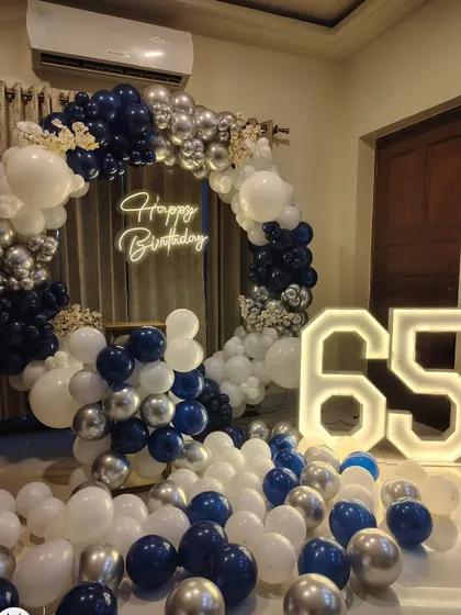 An elegant setup for a 65th birthday, featuring a navy, silver, and white balloon arch, a neon sign, and large marquee numbers.