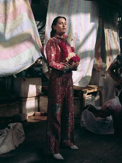 In this editorial shot, the model stands within the bustling flower market environment. The styling juxtaposes a structured, printed red suit against the chaotic, rustic background, creating a powerful high-low fashion statement.
