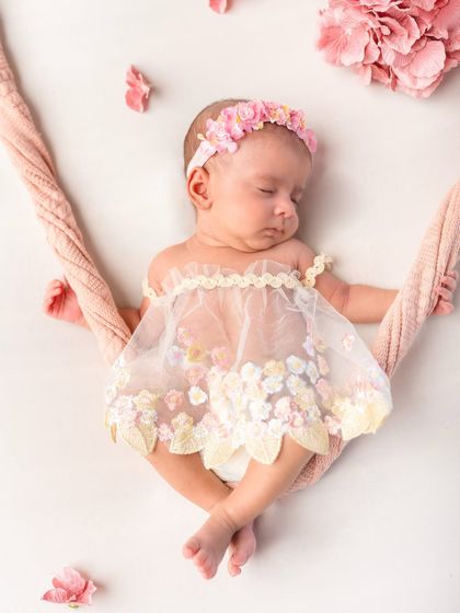 A newborn baby girl sleeps on a tiny floral swing, wearing a delicate dress. This is a beautiful and creative pose for a newborn session.