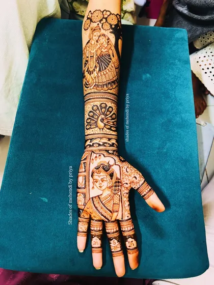 A full arm bridal mehendi with a bride portrait on the palm and a Radha Krishna figure on the forearm. The design is filled with details like peacocks and delicate finger patterns.