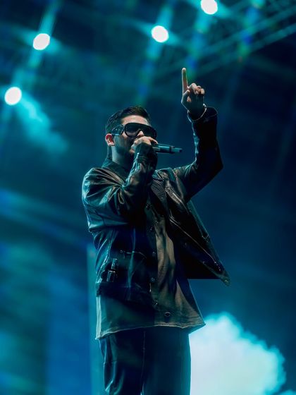 An artist points to the sky on a stage filled with smoke and light. This is a teaser image, designed to build anticipation for an upcoming major concert announcement.