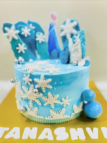 An icy blue Frozen cake featuring Elsa surrounded by a forest of edible snowflakes. The cool blue tones and shimmering details make it a magical winter wonderland.