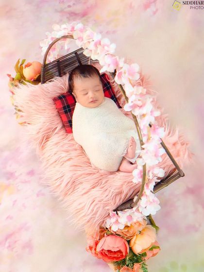A sleeping newborn, swaddled and resting in a hanging prop adorned with pink flowers. This artistic shot looks like a beautiful dream.