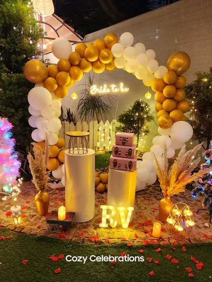 A festive "Bride to be" backdrop for a garden party. A circular balloon arch in gold and white frames a neon sign, with decorative Christmas trees and illuminated initials adding a personal and whimsical touch.