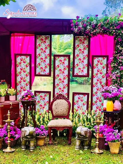 Here is an outdoor setup for a pre-wedding ceremony, featuring a backdrop of floral-printed panels and bright pink drapes. The area is decorated with fresh purple flowers, traditional brass lamps, and antique-style furniture for seating.