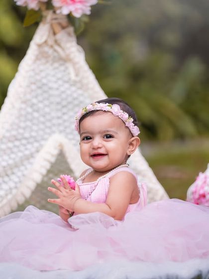 A happy baby girl enjoying her outdoor teepee setup, surrounded by pink flowers that complement her beautiful dress.