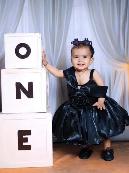 A stylish one-year-old in a black party dress, posing with 'ONE' blocks.