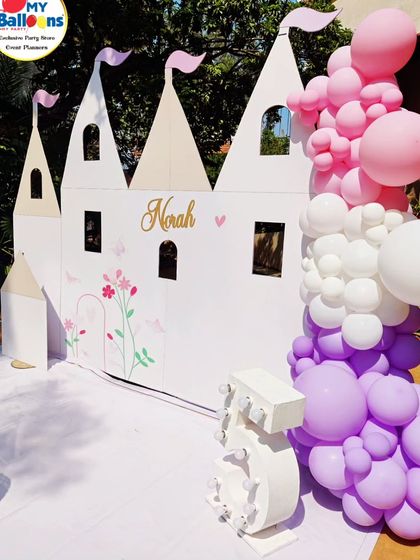 A life-sized castle backdrop for a fifth birthday. This simple yet elegant setup features a large white castle cutout, a single dessert plinth, and a purple and pink balloon garland.