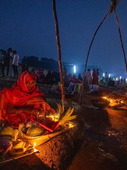 A woman performs rituals by the river's edge at night, surrounded by offerings and lamps. The scene is intimate and filled with a quiet, powerful spirituality.