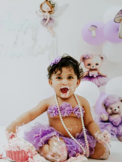 This little darling is all smiles, with cake on her face and hands. The soft purple and white balloon background completes the festive look.