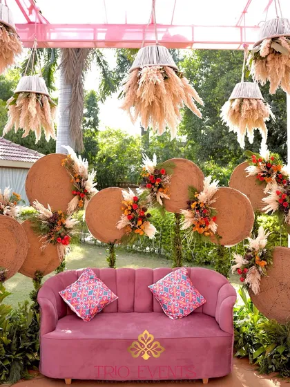 A full view of the boho-chic Mehendi seating area, complete with a plush pink sofa, woven disc backdrop, and hanging pampas grass chandeliers. This is a perfect spot for the bride to relax and enjoy her Mehendi.
