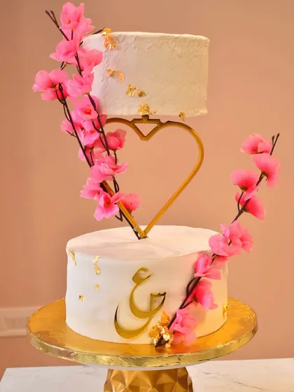 A modern and romantic floating engagement cake. This design uses a custom heart-shaped separator to create the illusion of a floating top tier, decorated with delicate pink cherry blossoms and personalized gold calligraphy.