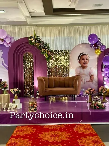 We designed this royal princess theme for a first birthday, using a rich purple color palette. The stage includes multiple arched backdrops, a miniature sofa for photos, a light-up number '1', and a princess carriage prop, all set on a purple carpeted stage.