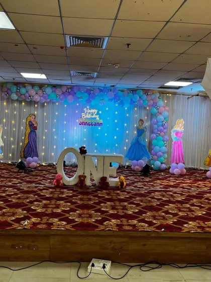 A wide-angle view of a princess-themed first birthday in a large hall. The L-shaped balloon arch and fairy lights create a magical backdrop for the 'ONE' table and character cutouts.