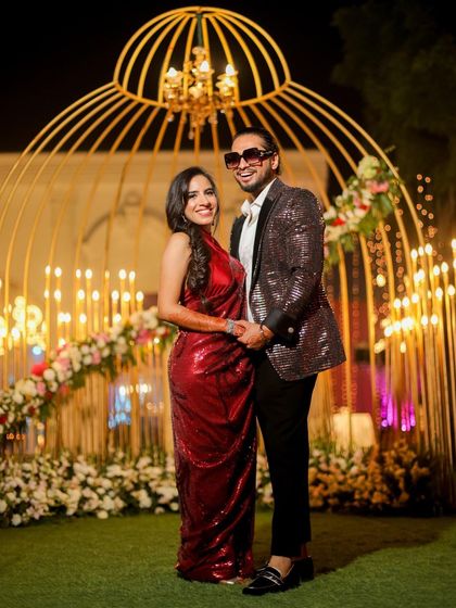 A classic portrait of a newly engaged couple. They stand before a stunning golden birdcage-like decor piece, holding hands and smiling, perfectly capturing the elegance of their engagement party.