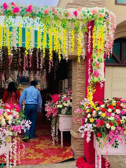 A colorful and festive entrance for a home-based wedding event. The walkway is decorated with hanging floral strings and large bouquets, creating a welcoming passage for guests.