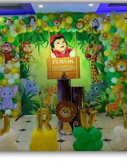 A jungle-themed first birthday stage with a photo backdrop and the name 'PURVIK' in foil balloons.