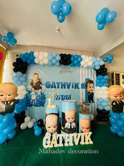 A Boss Baby themed first birthday party in a hall. This setup uses a large backdrop with character graphics, multiple Boss Baby cutouts on balloon pillars, and the birthday boy's name in wooden letters.