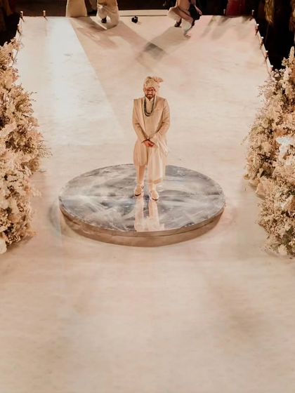 The groom stands on a mirrored platform at the start of the aisle, awaiting his bride. The aisle is lined with what looks like a forest of white florals, creating a dramatic and ethereal path.