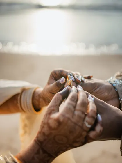 An intimate, close-up photo of the ring exchange during a beach engagement ceremony, with the sparkling ocean in the background.