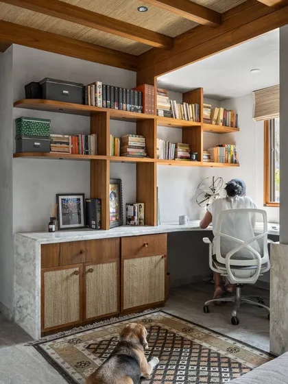 A home office designed for focus and comfort. The custom wooden desk and shelving unit provide ample storage, while the material palette of concrete, wood, and cane creates a calming work environment.