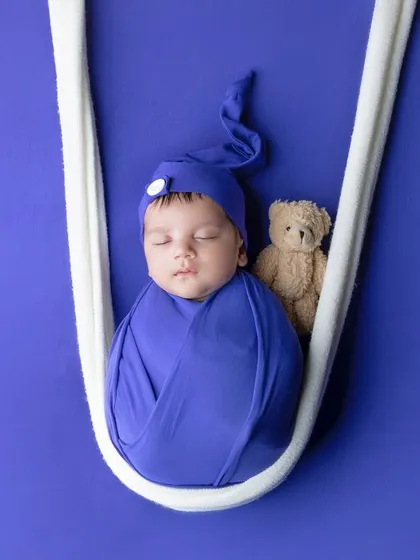 A baby boy in a royal blue wrap and hat, sleeping in a white sling with his teddy.