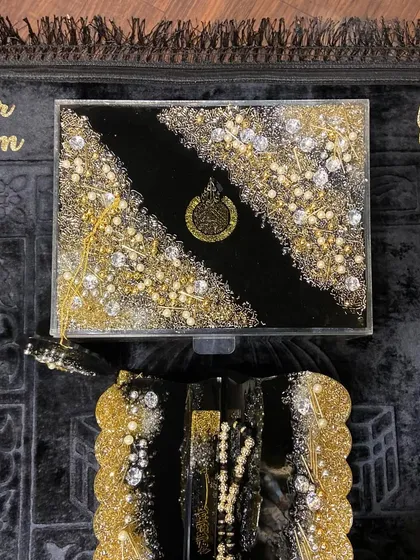 A top-down view of my trending black and gold Islamic gift set. This luxurious collection includes a personalized prayer mat, a resin Quran box, and a matching Quran holder, all heavily embellished for a grand look.