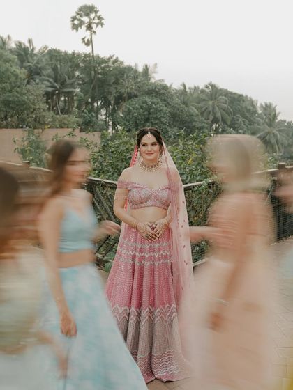 A creative shot of the bride in her pink lehenga, with her bridesmaids blurred in motion around her. The focus remains on her calm and central presence amidst the happy chaos.
