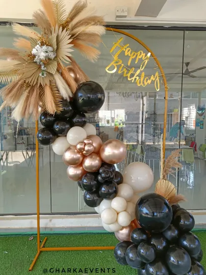 A boho-chic birthday arch with black, white, and rose gold balloons. The arrangement is beautifully accented with dried pampas grass and palm leaves for a natural, trendy look.